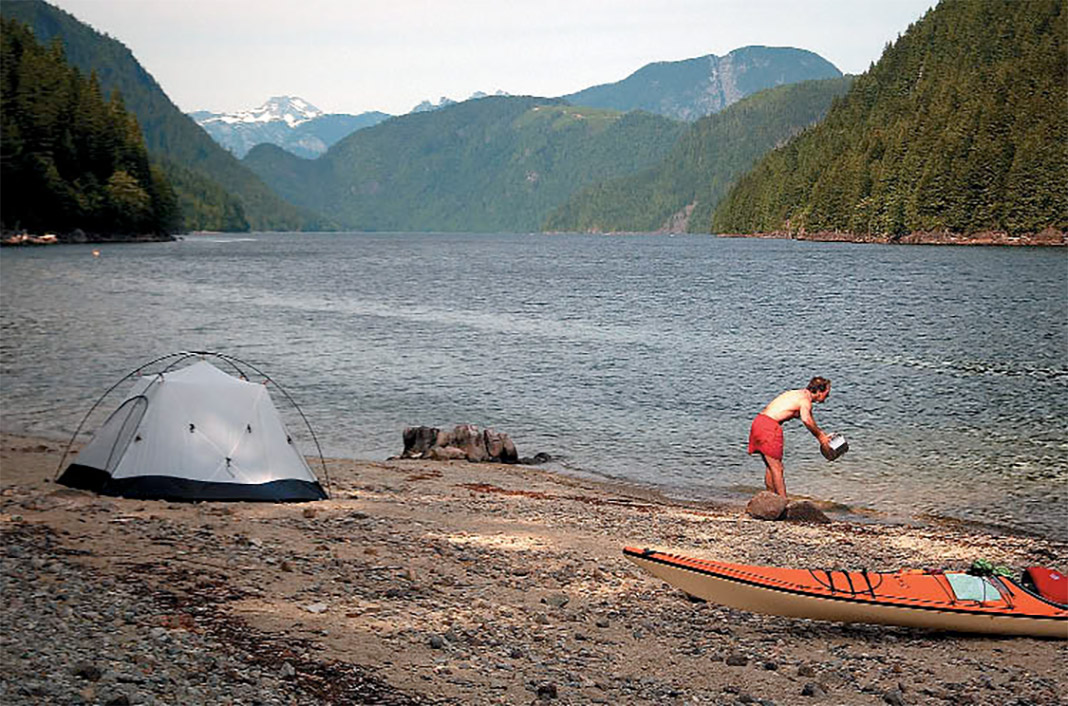 Man standing at edge of lake with kayak beside him and tent on beach.