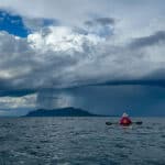 A sea kayaker sits on the water looking at a storm on the horizon