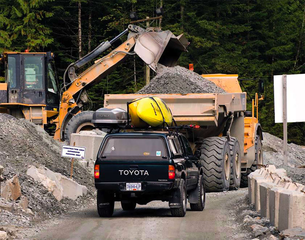 Toyota truck with yellow kayaks on roof waiting behind dump truck full of gravel