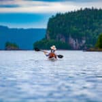 woman paddling away from the camera along a rocky, wild shore