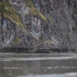 The Rapids Are Real, The River Isn’t people paddle a canoe in front of a large grey rock face