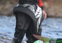Person standing beside whitewater kayak wearing sprayskirt.