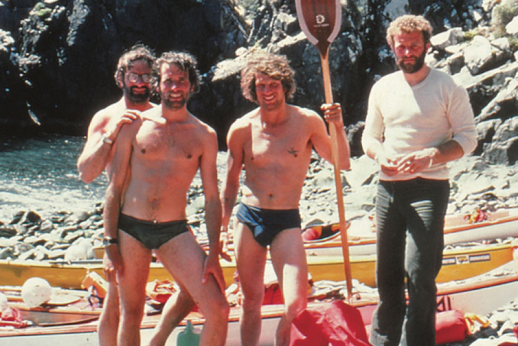 Four men stand in front of a group of Nordkapp kayaks, one holding a paddle