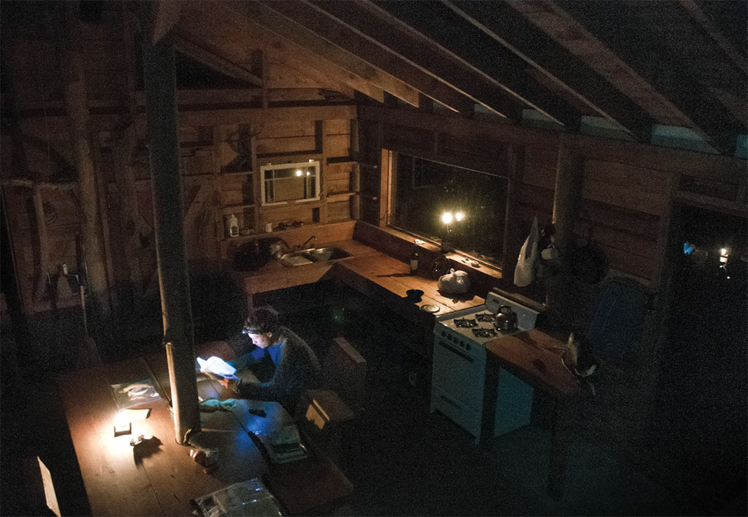 Man sitting at table in a cabin reading a book by headlamp.
