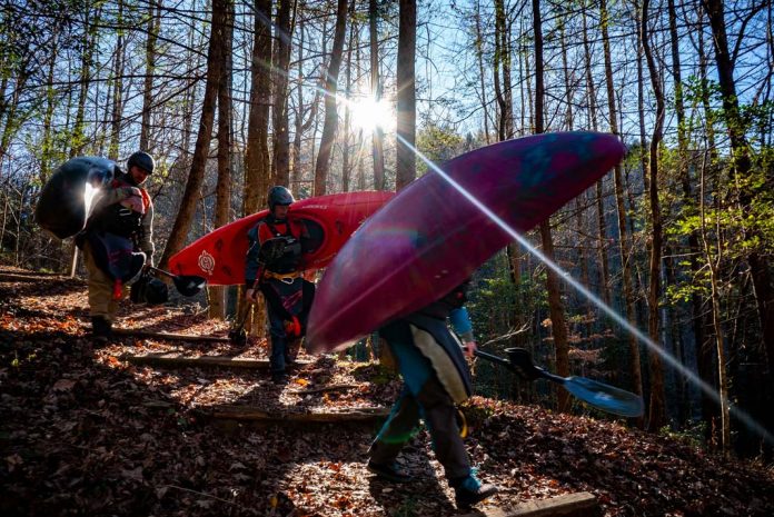 Three people carrying whitewater kayaks through the woods