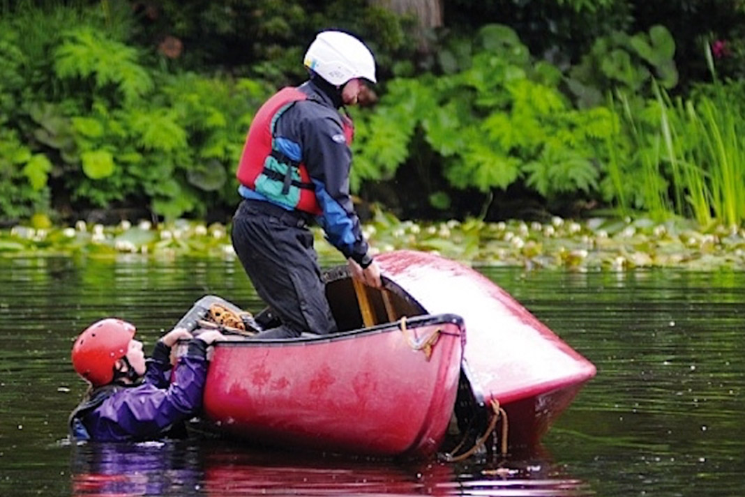 One person standing in canoe lifting a swamped canoe, another person holding the gunwales of the stable canoe.
