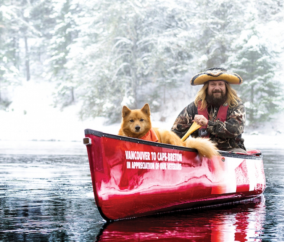 Man paddling a canoe with dog in bow, snow on trees in background.