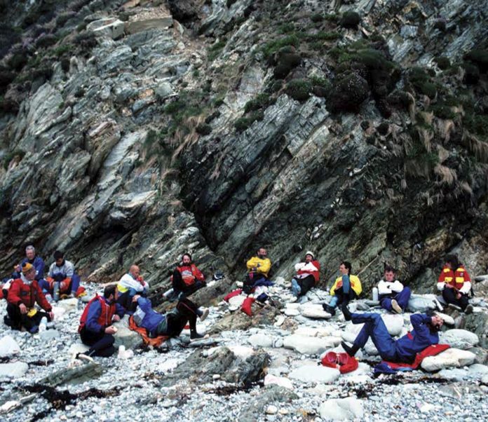 THE LATE FRANK GOODMAN AND A GROUP OF FRIENDS, BREAK FOR LUNCH ON A GRAVEL BEACH ON ANGLESEY ISLAND, WALES.| Photo by: Stan Chladek