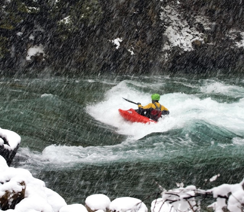 A whitewater kayaker surfs a wave on a river in winter. 