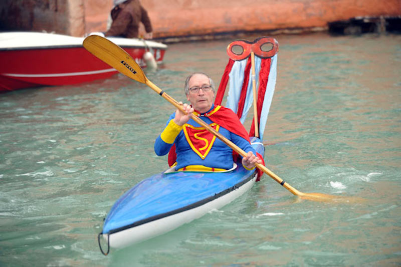 A man in a Superman costume paddling a blue kayak. 