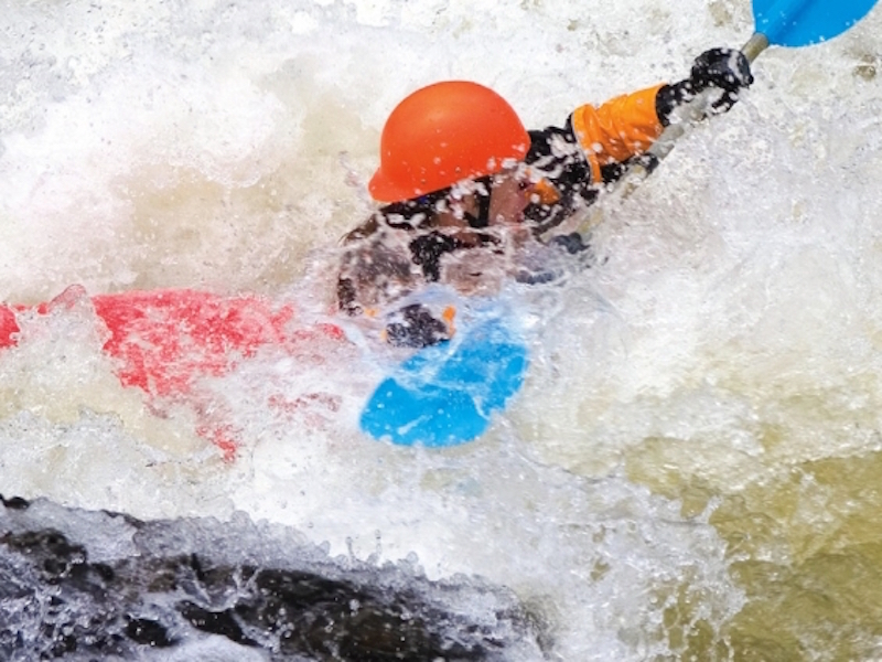 A kayaker in an orange helmet is stuck in a hole on a whitewater river. 