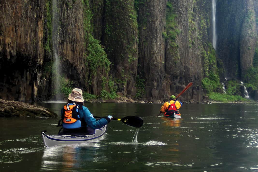 two people paddling past large cliffs with waterfalls, perhaps in search of a site for free camping