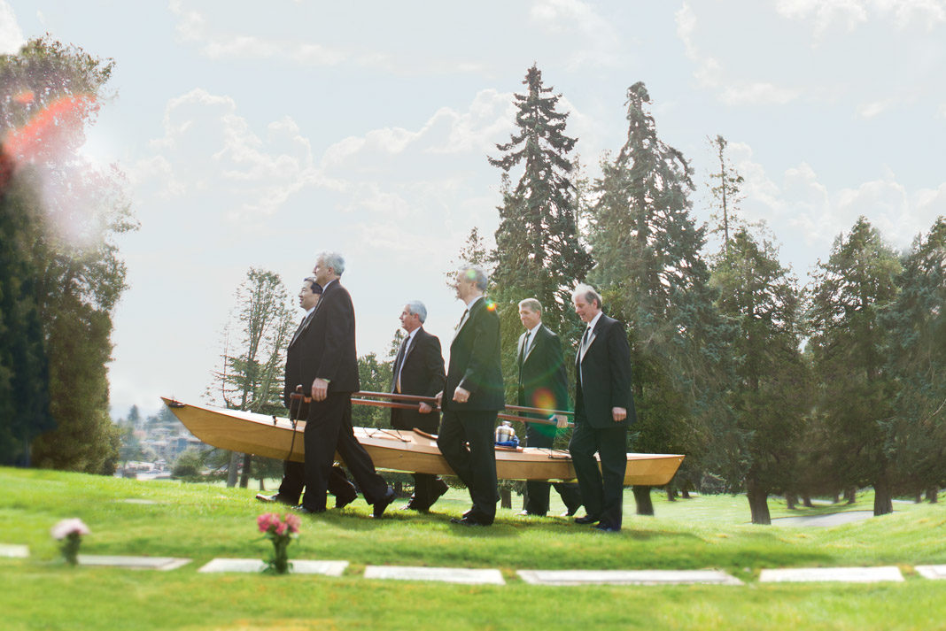 6 men in tuxedos carrying a wooden kayak with an urn