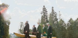 6 men in tuxedos carrying a wooden kayak with an urn