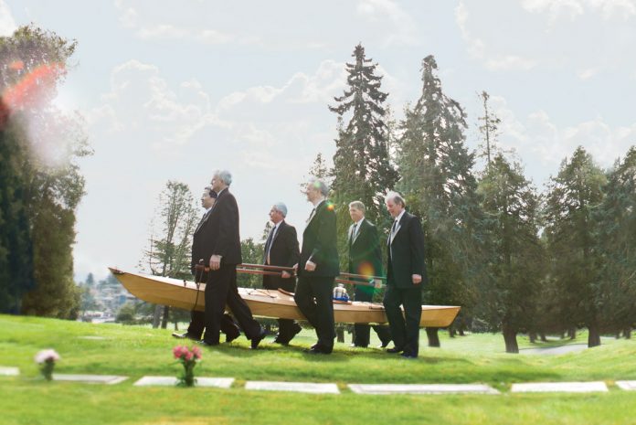 6 men in tuxedos carrying a wooden kayak with an urn