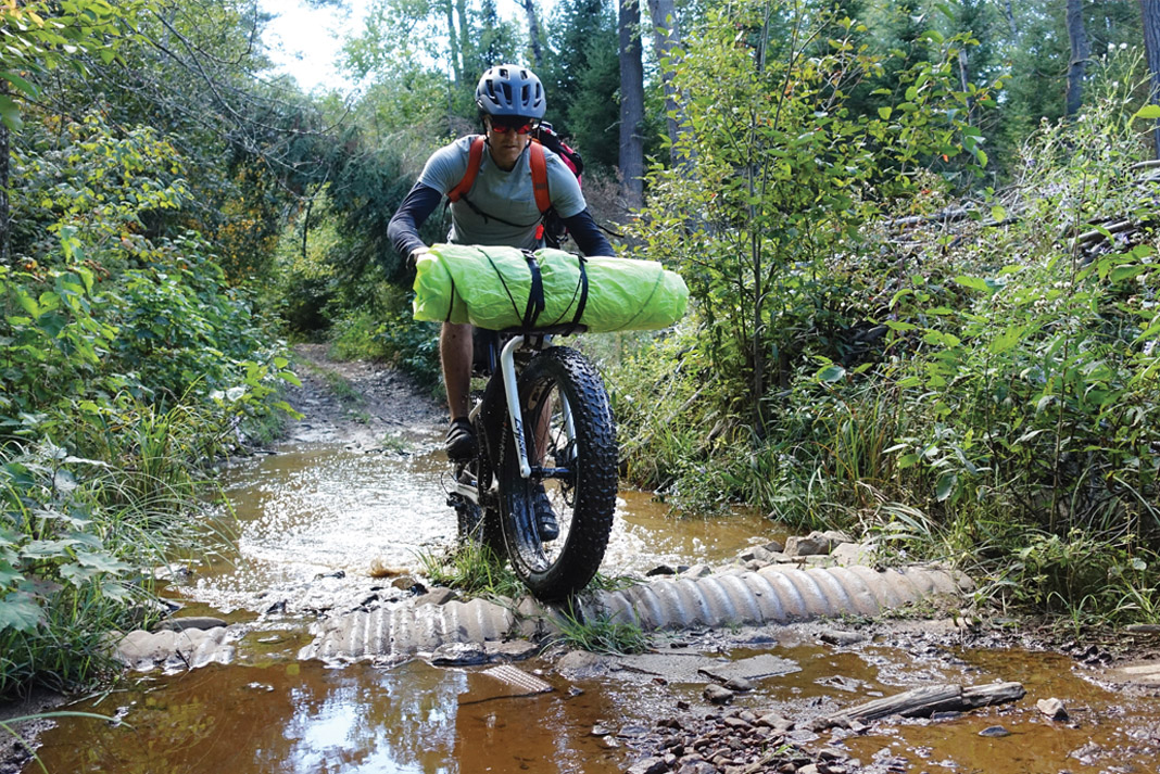 man riding a fat bike through puddles while carrying a rolled-up packraft