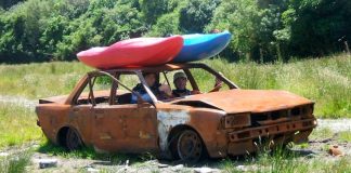 two men in a broken down car with two kayaks strapped to the hood of the car