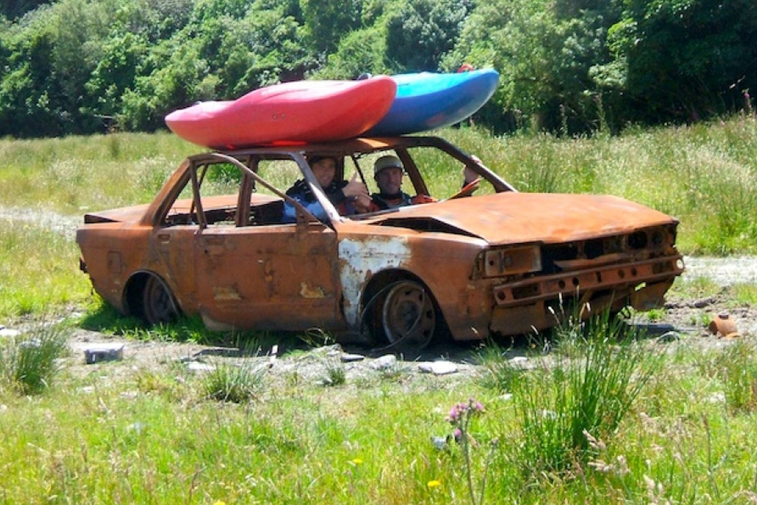 two men in a broken down car with two kayaks strapped to the hood of the car