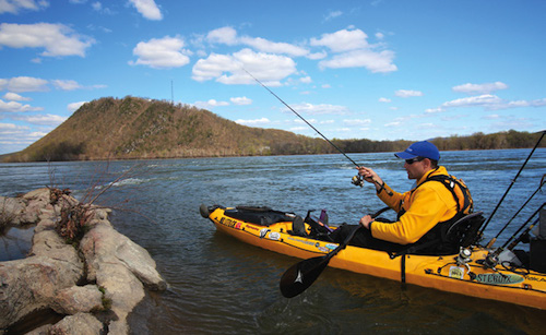 A fishing kayaker casts behind a rock in a river.