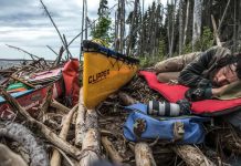 man sleeping next to his canoe surrounded by paddling and photography gear