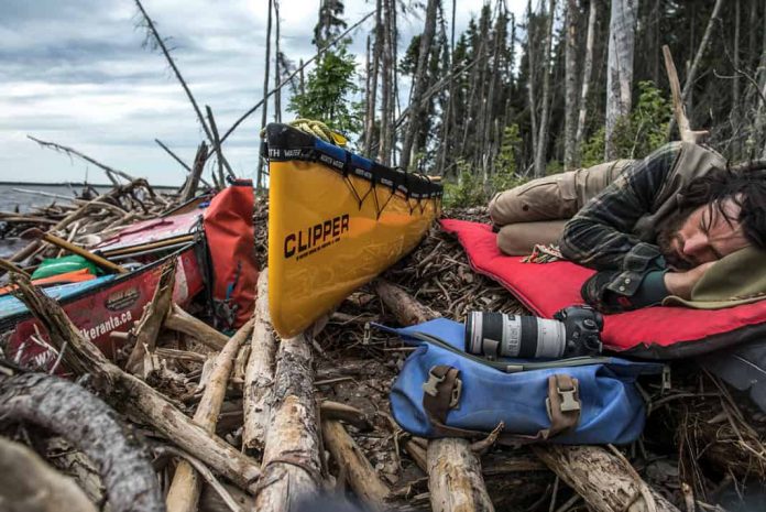man sleeping next to his canoe surrounded by paddling and photography gear