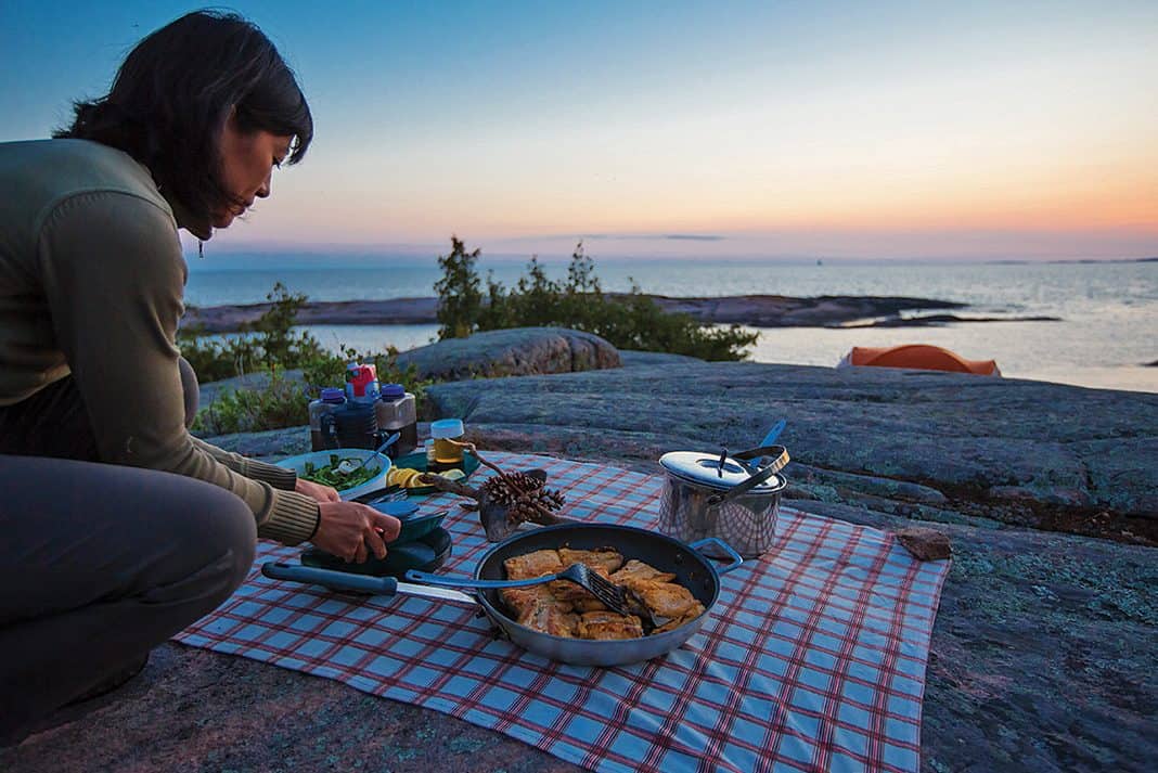 A camper prepares dinner on the rocky edge of a large body of water, late in the evening