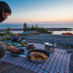 How To Eat Fresh On Your Camping Trips A camper prepares dinner on the rocky edge of a large body of water, late in the evening