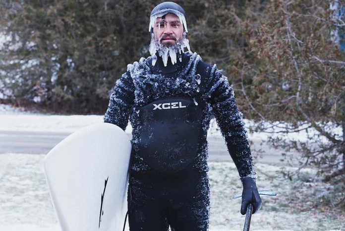 man standing with his paddleboard in the winter covered with icicles on his face