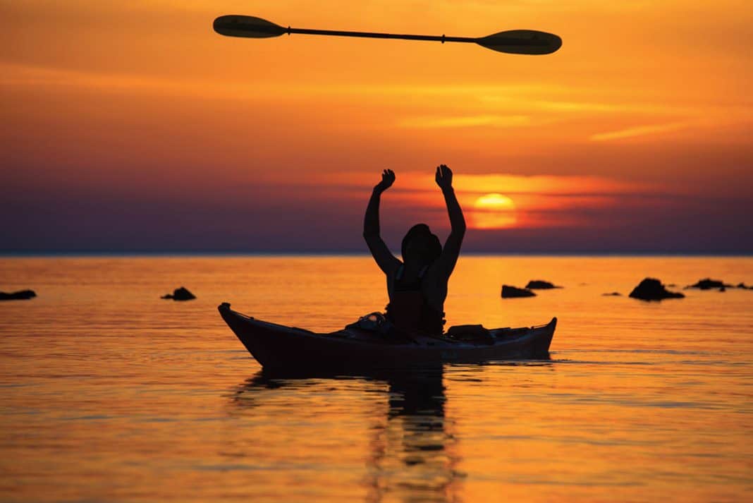 a kayaker tossing up their kayak paddle with the sun setting in the background on calm waters