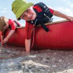 10 Tips To Survive On A Family Canoe Trip two kids in a canoe that has been beached playing with the water underneath them