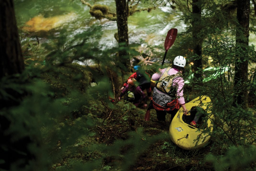 3 kayaker's dragging their boats through a forest with their kayaks and kayak paddles looking at whitewater