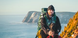 Man wearing pack kneeling behind dog, also wearing a pack, with ocean and cliffs in background.