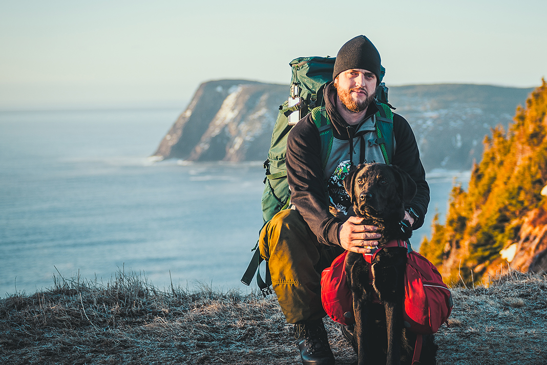 Man wearing hiking pack kneeling behind dog, also wearing a pack, with cliffs and ocean in background.