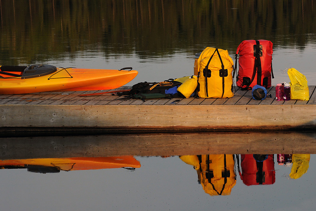 Many drybags sitting on a dock with a kayak in the water