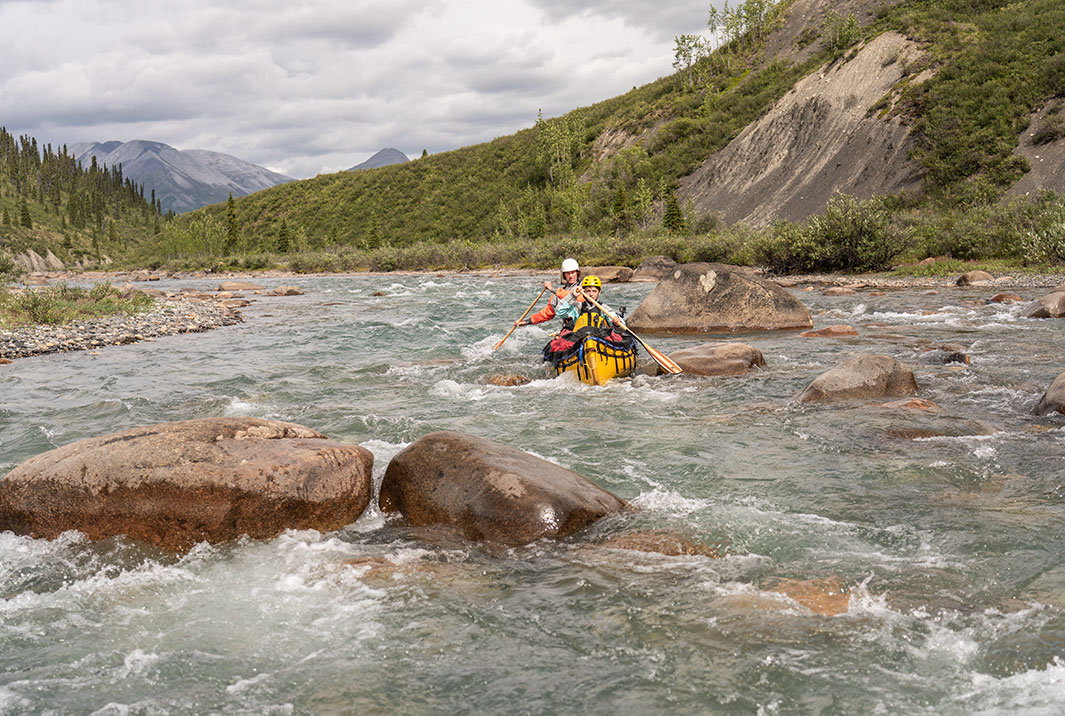 Two people in a yellow canoe going through boulder-strewn rapids
