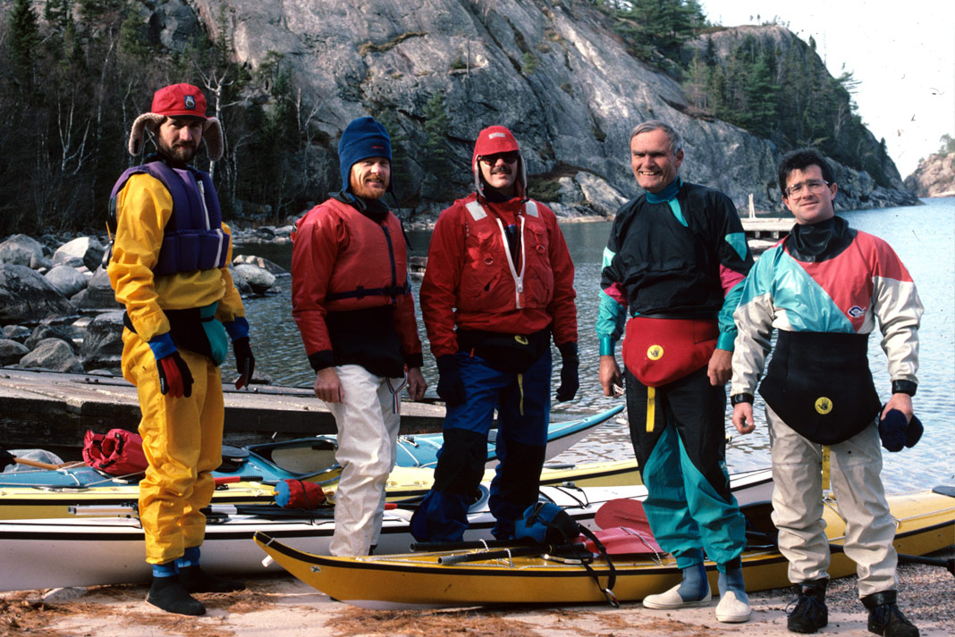Five men standing beside a lake with sea kayaks at their feet.