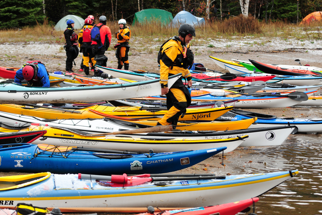 Tons of sea kayaks on a beach, with one kayaker wearing a drysuit trying to launch.