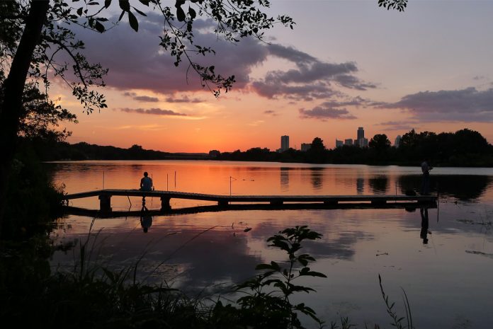 A person sitting on a dock silhouetted by sunset across the water