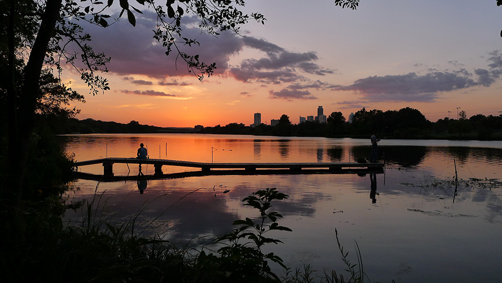 A person sitting on a dock silhouetted by sunset across the water