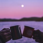 Tim’s Wilderness Rules Two hands clink camping mugs in front of a lake during a pink and purple sunset