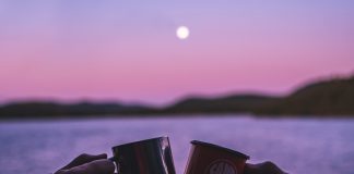 Two hands clink camping mugs in front of a lake during a pink and purple sunset