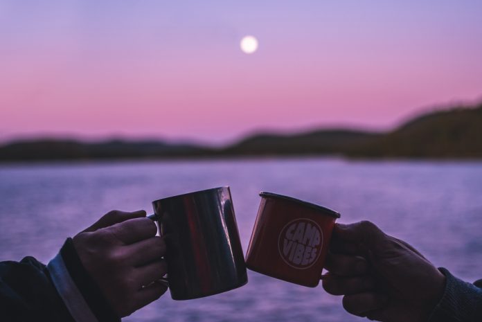 Two hands clink camping mugs in front of a lake during a pink and purple sunset