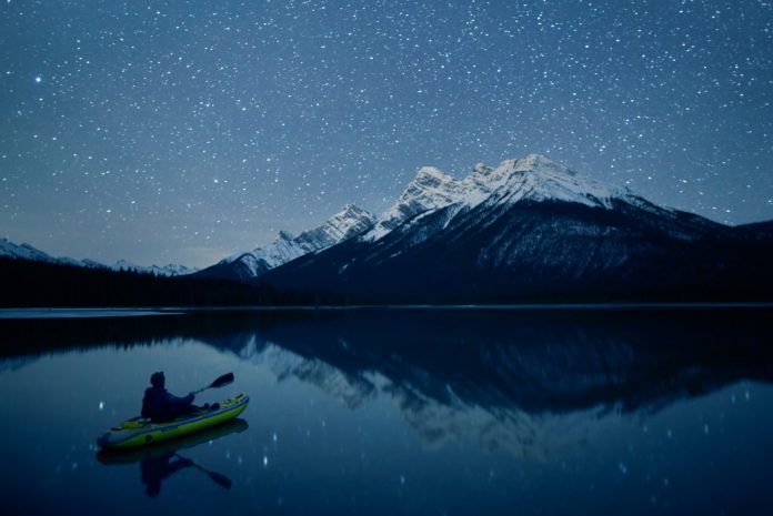 A paddler kayaks towards a mountain under a starry sky at night.