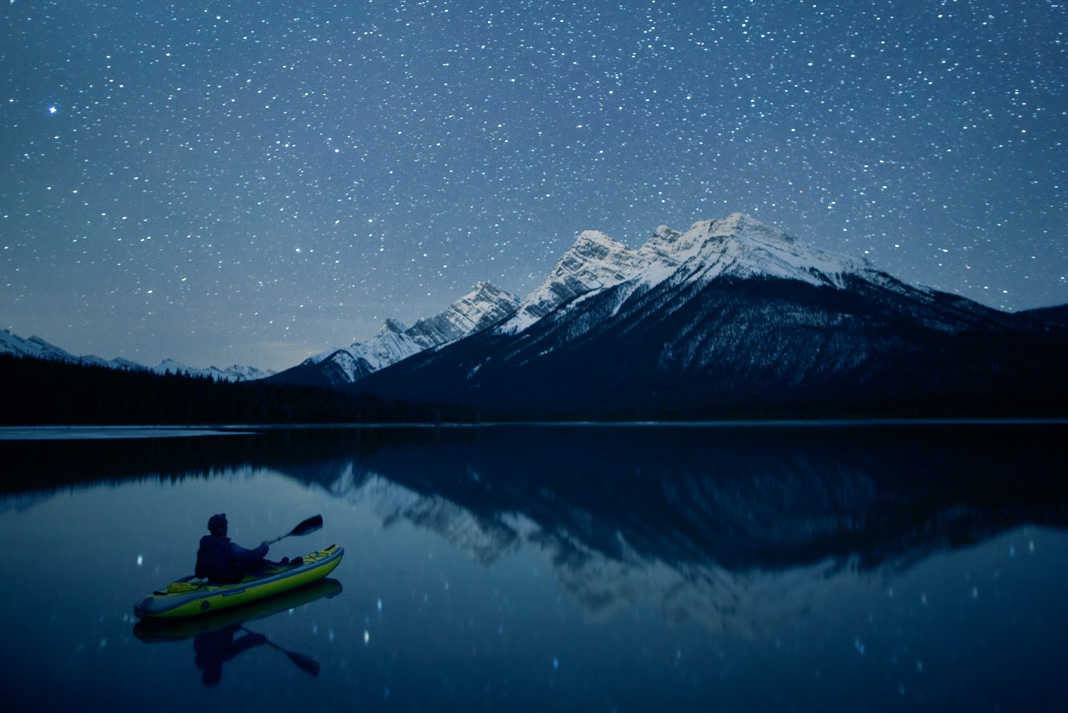 A paddler kayaks towards a mountain under a starry sky at night.