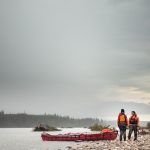 The Paddler’s Greatest Debates Two canoeists stand at the side of a lake against a grey sky