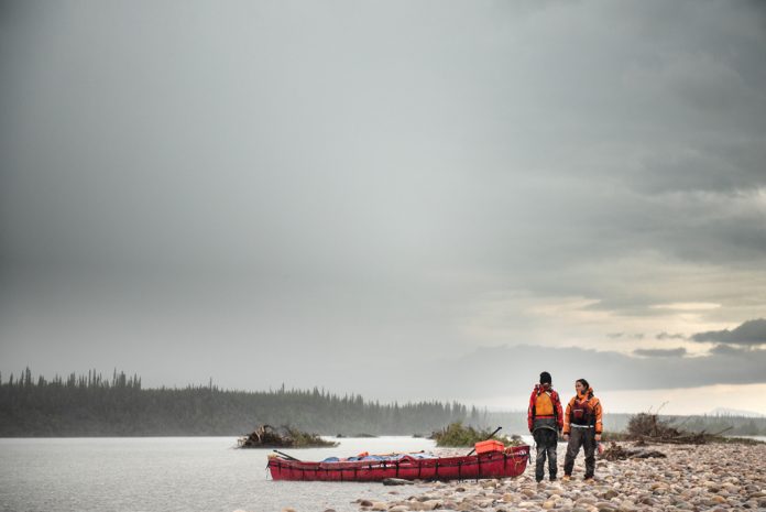 Two canoeists stand at the side of a lake against a grey sky