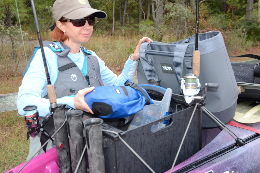 Woman standing beside fishing kayak with cooler, drybag, and other gear.