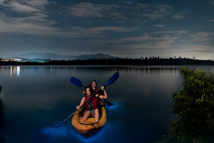 Girls kayaking on a bioluminescent bay under a cloudy night sky