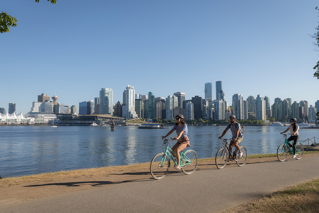 Three people cycling on paved path next to water with skyscrapers in background.