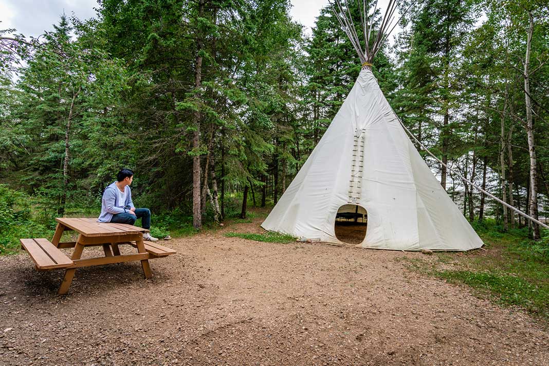 A man sits on a picnic table outside a tipi while camping near Edmonton